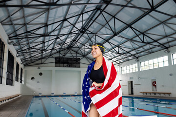 Young biracial female swimmer standing by pool indoors, wrapped in American flag, copy space