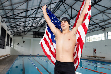 Caucasian young male swimmer holding American flag, standing by pool indoors, celebrating