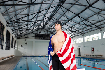 Caucasian young male swimmer standing by pool indoors, draped in American flag, copy space