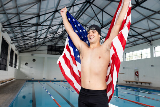 Caucasian young male swimmer holding American flag, standing by pool indoors, celebrating - Powered by Adobe