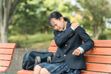 登校・下校中に肩を痛めて公園のベンチに座る中学生・高校生の女子生徒
