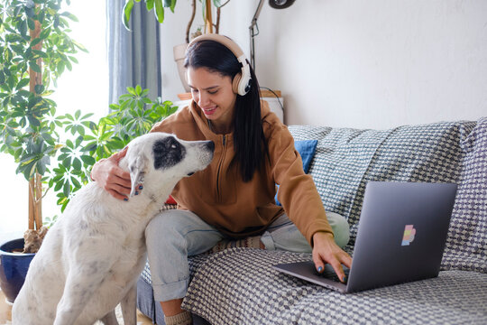 Woman with headphones using laptop on sofa bonding with dog 