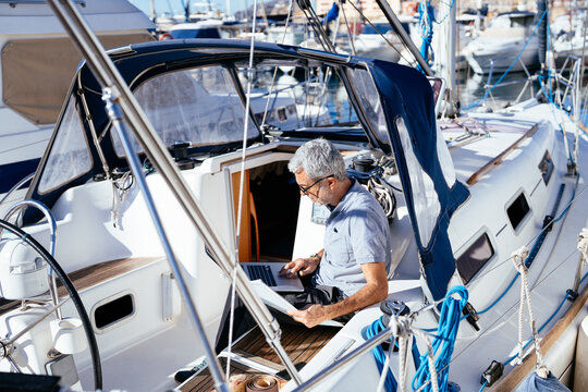 Senior man checking information on laptop while holding papers on boat