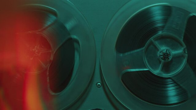 Retro-styled open reel tape recorder with rotating spools in dark studio with blue neon light. Zoom off, close-up shot