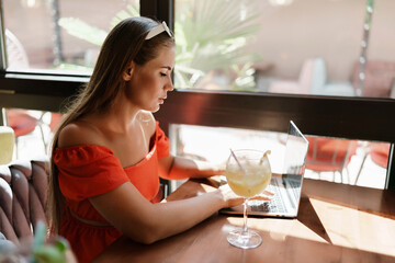 Woman works on laptop in a sunny cafe, glass of juice nearby. The setting is modern, with natural light illuminating the space. The image captures a moment of focused work.