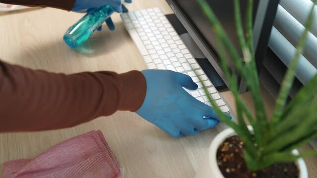 A person is cleaning a computer keyboard with a blue glove on. A potted plant is on the desk next to the keyboard