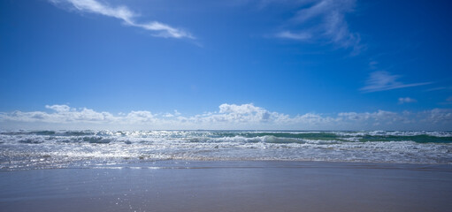 Wild surf, waves, sea and sand in a panoramic seascape with blue cloudy sky and brilliant sunshine...