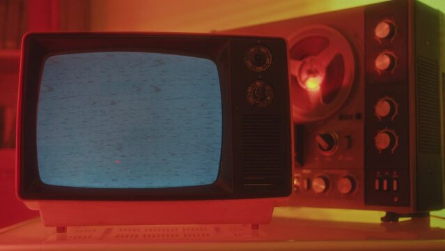Old-fashioned analog TV displaying static noise and reel-to-reel tape recorder in a dark studio with red neon light. Close-up shot, no people