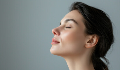 Fototapeta premium A profile view of a serene woman with eyes closed, her expression one of peace and introspection, set against a soft grey backdrop.