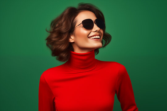 A woman's joyful laughter captures a moment of timeless elegance, her chic red turtleneck complemented by classic round sunglasses, against a deep green backdrop.