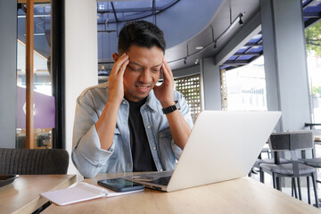 Asian man get pressure Tired headache, having Neck Pain, using laptop, mobile phone in coffee shop,...