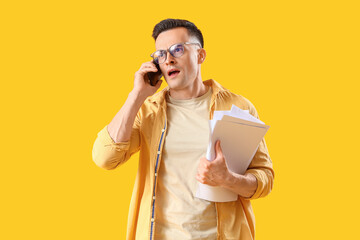 Stressed young man with papers talking by mobile phone on yellow background. Deadline concept
