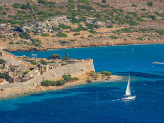 Yacht cruising by Spinalonga island and peninsula behind (Plaka, Crete, Greece) - obrazy, fototapety, plakaty