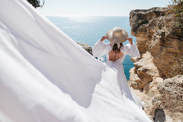 A woman in a white dress is standing on a rocky cliff overlooking the ocean. She is wearing a straw...