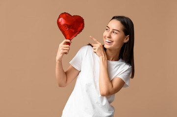 Beautiful young woman pointing at heart-shaped air balloon on beige background