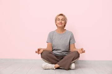 Portrait of meditating mature woman sitting on floor against pink wall