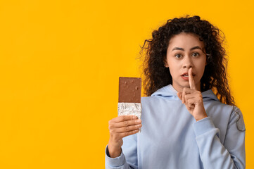 Beautiful young African-American woman with sweet chocolate bar showing silence gesture on yellow...
