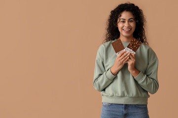 Happy young African-American woman with sweet chocolate bars on brown background