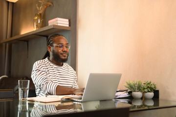 Young African American man in eyeglasses sitting at table, working on laptop at home