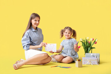Naklejka premium Mom with her little daughter sitting on floor with bouquet of tulips and greeting card on yellow background. Mothers Day concept