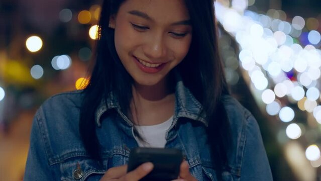 Beautiful young Asian woman standing in city street at night using mobile smartphone. Happy woman standing and typing messages on her mobile phone