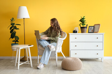 Beautiful young woman using laptop in wicker armchair at home