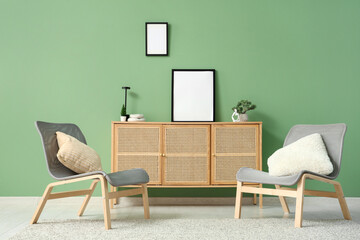 Stylish living room with cozy armchairs, chest of drawers and blank picture frames on green wall