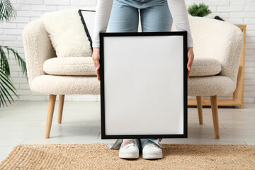 Young woman with blank picture frame near sofa in living room