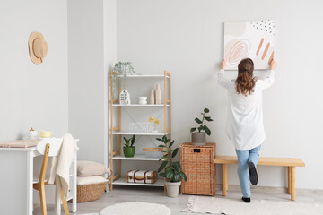 Young woman hanging picture on white wall in room
