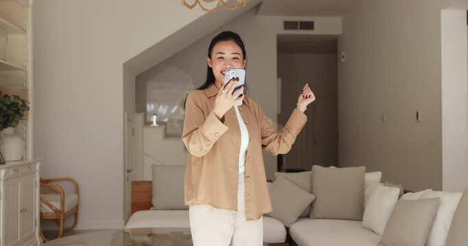 Untroubled Asian woman standing in living room with smartphone watch music video and dancing, spend pastime at home, move to favourite song looks happy and carefree. Tech, hobby, social media platform