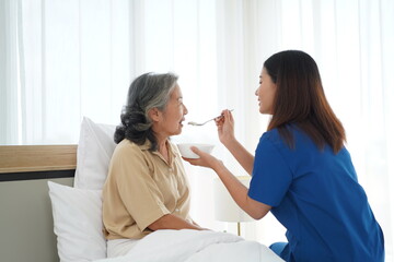 Asian young caregiver female feeding aged woman on bed at home