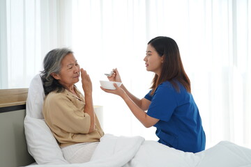 Asian young caregiver female feeding aged woman on bed at home