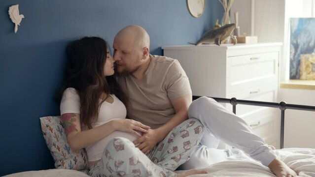 Mid adult couple expecting baby while caress his wife. Happy father hands on expecting mother's baby bump while embracing her on bed. Family, parenthood. Young parents preparing to have child