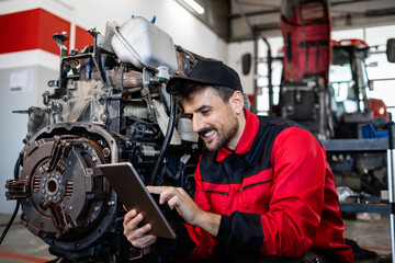 Serviceman checking spare parts inventory on tablet computer and repairing tractor engine.