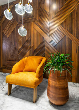Modern orange armchair, beside tall wooden planter with green bushes, and contemporary tall glass chandelier, in a hall with decorated wood cladding wall, and white marble floor