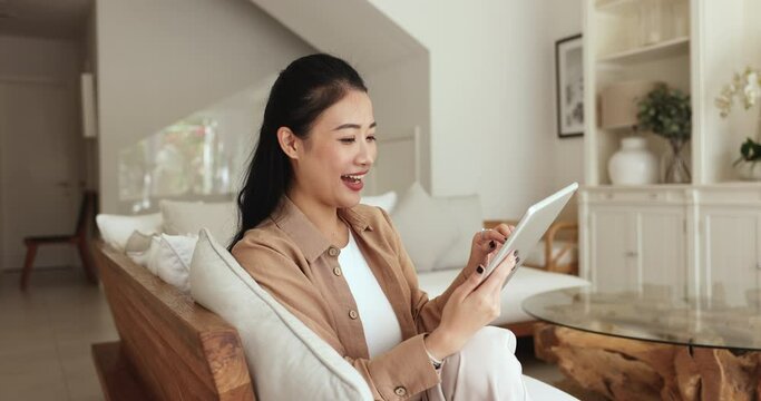 Asian woman spend free time on internet, resting seated on sofa with digital tablet, chatting on-line with family or boyfriend, using e-dating services on weekend at home. Wireless tech usage, leisure
