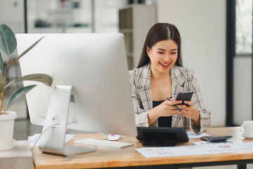 Joyful Asian businesswoman using smartphone at office workstation, with a desktop computer and financial reports on her desk.