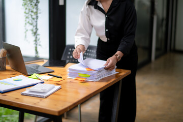 Focused businesswoman in a two-tone shirt sorting through stacks of paperwork at a wooden desk in an office environment.