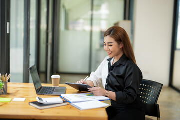 Professional Asian businesswoman typing on laptop at a wooden desk with paperwork and greenery in a bright office setting.
