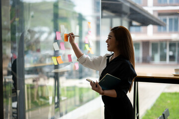 Smiling Asian businesswoman using colorful sticky notes for brainstorming on a glass wall in a...