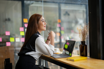 Professional Asian businesswoman typing on laptop at a wooden desk with paperwork and greenery in a bright office setting.