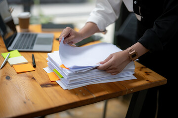 Focused businesswoman in a two-tone shirt sorting through stacks of paperwork at a wooden desk in an office environment.