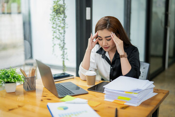 Pensive Asian businesswoman reading a report, with a laptop and paperwork on her desk in a bright office space.