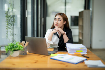 Pensive Asian businesswoman reading a report, with a laptop and paperwork on her desk in a bright office space.