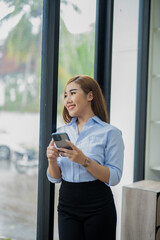 Smiling young Asian businesswoman using her smartphone by the window in a well-lit office, dressed in smart casual attire.