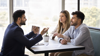 Positive married couple consulting financial expert, real estate agent at meeting, listening to young Indian business consultant in formal suit, sitting at table with paper agreement