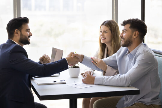 Happy young diverse couple shaking hands with Indian consultant, realtor, broker. Successful real estate agent, financial specialist, legal advisor giving handshake to satisfied clients