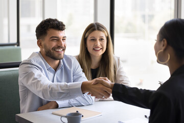 Cheerful young couple shaking hands with African American lawyer, broker, consultant, real estate agent, thanking for consultation. Positive business partners giving handshake at meeting, smiling