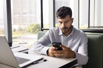 Serious young Arab businessman typing on smartphone on job chat, reading text message, using mobile phone for online communication at co-working workplace with laptop