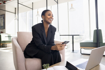 Happy attractive young African American business lady using Internet technology for job in co-working space, sitting in armchair at laptop, holding mobile phone, looking away, smiling